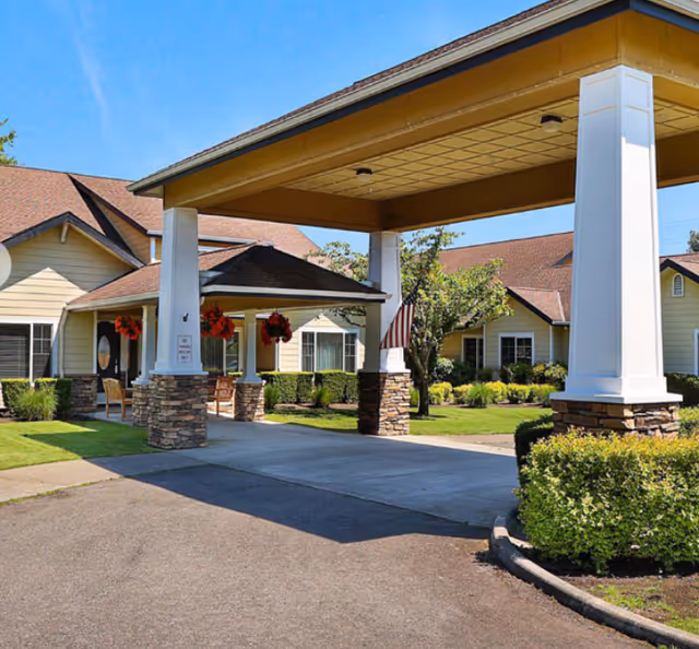 Entrance area of a senior living facility with a covered driveway supported by white columns with stone bases. The building has beige siding and brown shingled roofs, with hanging flower baskets and an American flag near the entrance. Well-maintained landscaping with green bushes and trees surrounds the area under a clear blue sky.