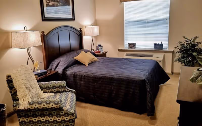 A furnished bedroom with a double bed, two bedside lamps, an upholstered armchair, a window and potted plants.