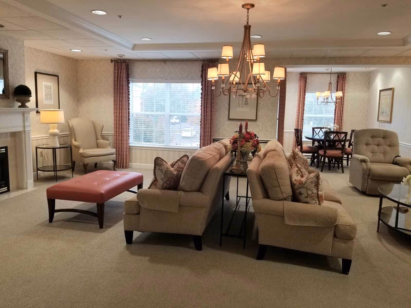 A cozy living room area in a senior living facility featuring two beige sofas with patterned pillows facing each other, a pink upholstered ottoman, a beige armchair, a fireplace with a lamp and decorative plant on the mantel, and a dining area with a table and chairs in the background. The room has beige carpeting, patterned wallpaper, and large windows with red curtains letting in natural light.