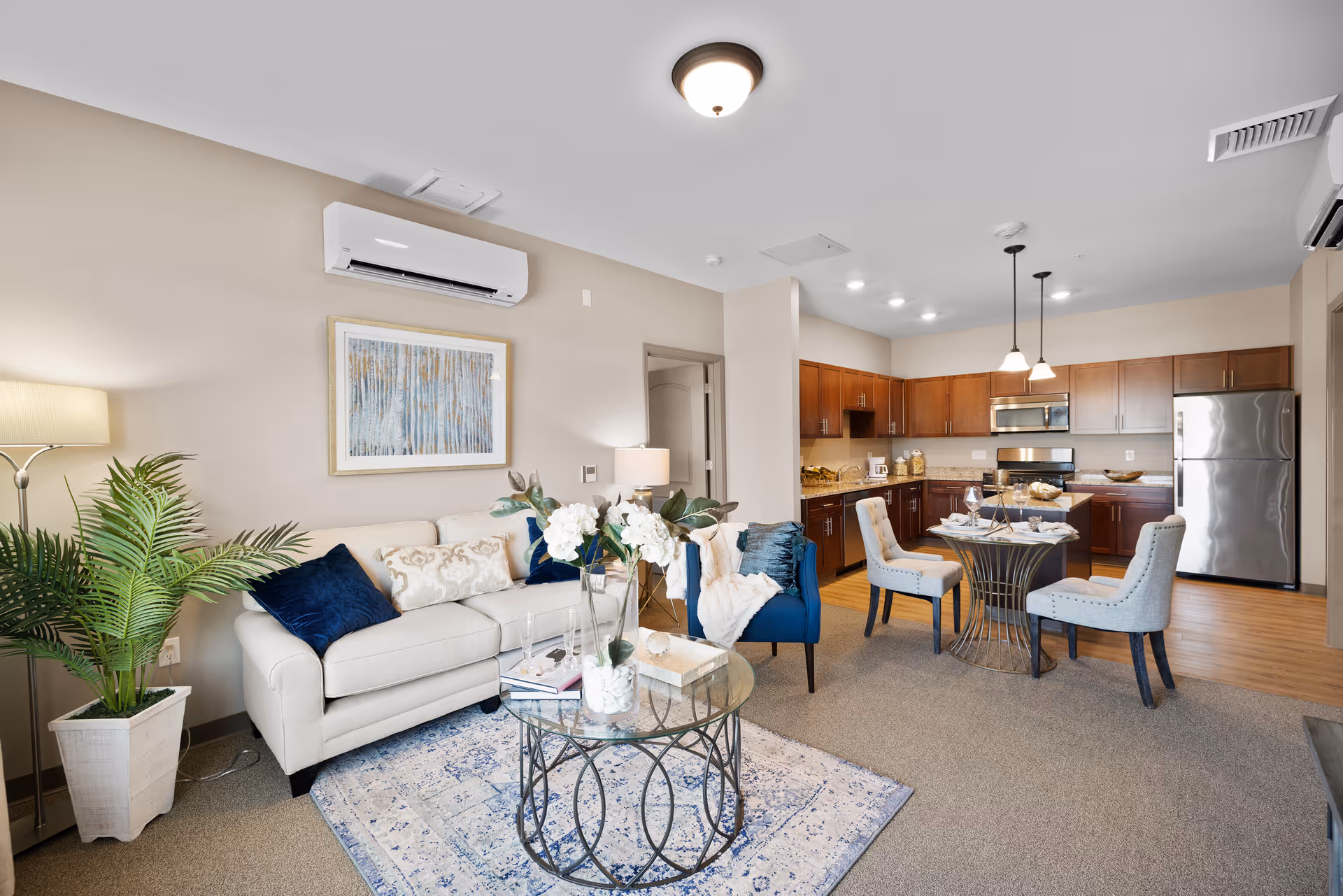 A modern open-concept living room and kitchen area in a senior living facility. The living room features a white sofa with blue and beige pillows, a blue armchair, a glass coffee table with decorative items, a floor lamp, and a potted plant. The kitchen has wooden cabinets, stainless steel appliances including a refrigerator and microwave, and a small round dining table with four upholstered chairs set for a meal. The walls are painted beige, and the floor is a combination of carpet and wood.