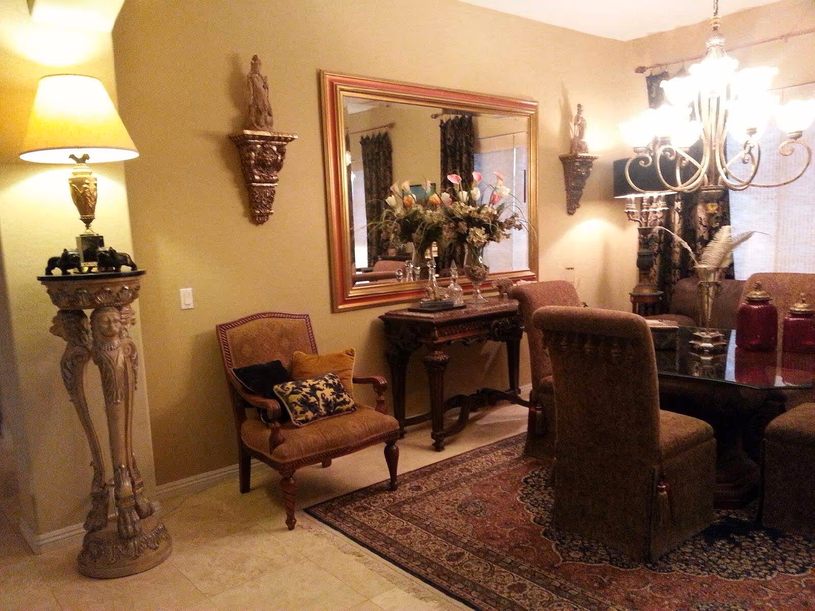 A formal dining room with ornate chairs around a glass table, a chandelier, large mirror, decorative pedestal and floral centerpiece.