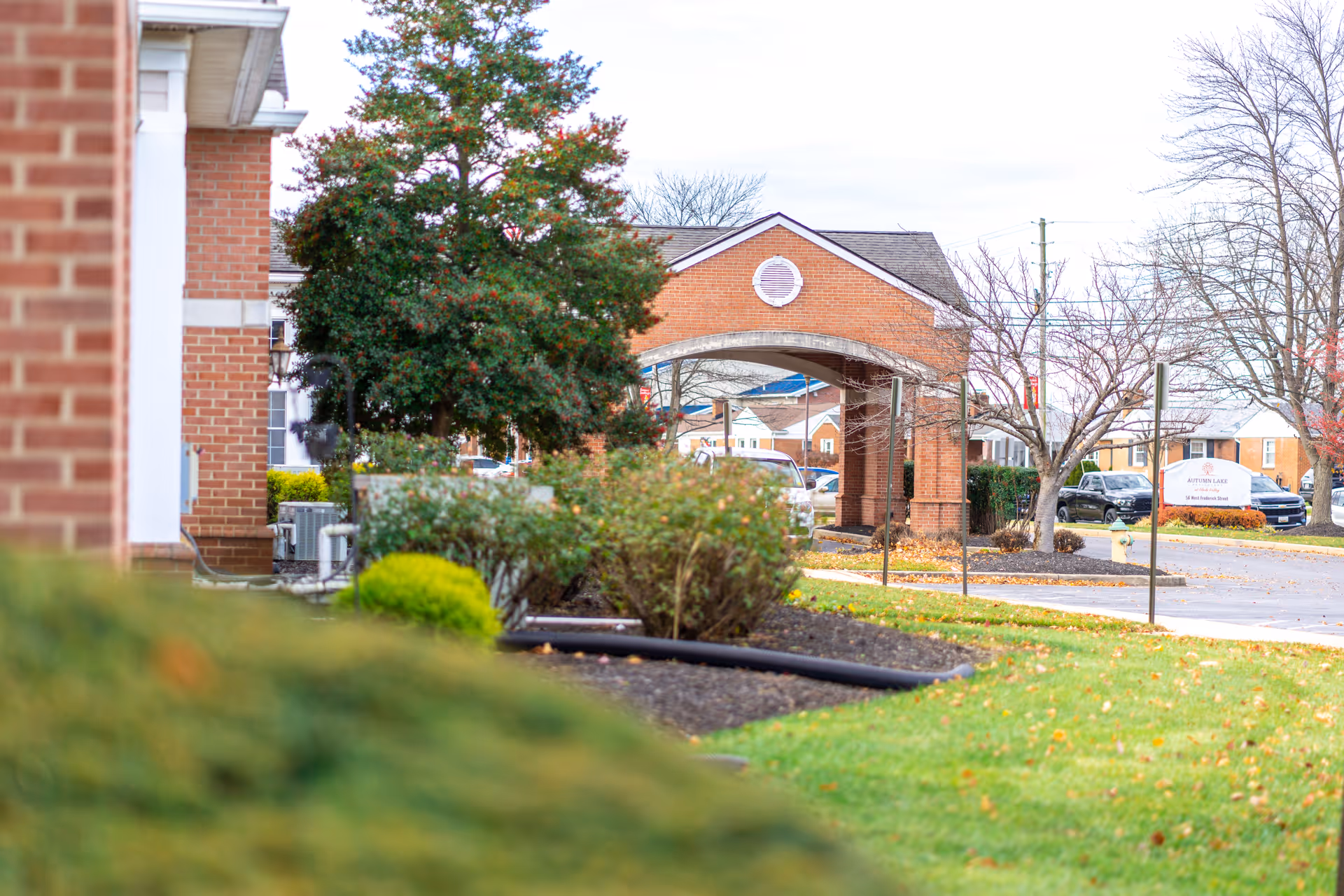 View of the exterior of Autumn Lake Healthcare at Glade Valley showing a brick building with a covered entrance, landscaped bushes, trees, and a parking area with cars in the background.
