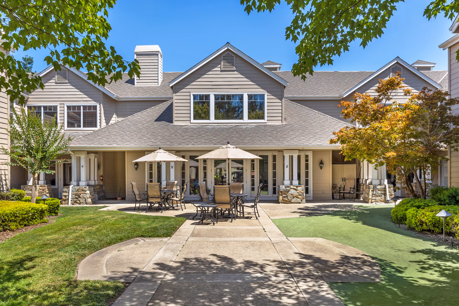 Outdoor courtyard with patio tables and umbrellas in front of a two-story residential building surrounded by landscaping.