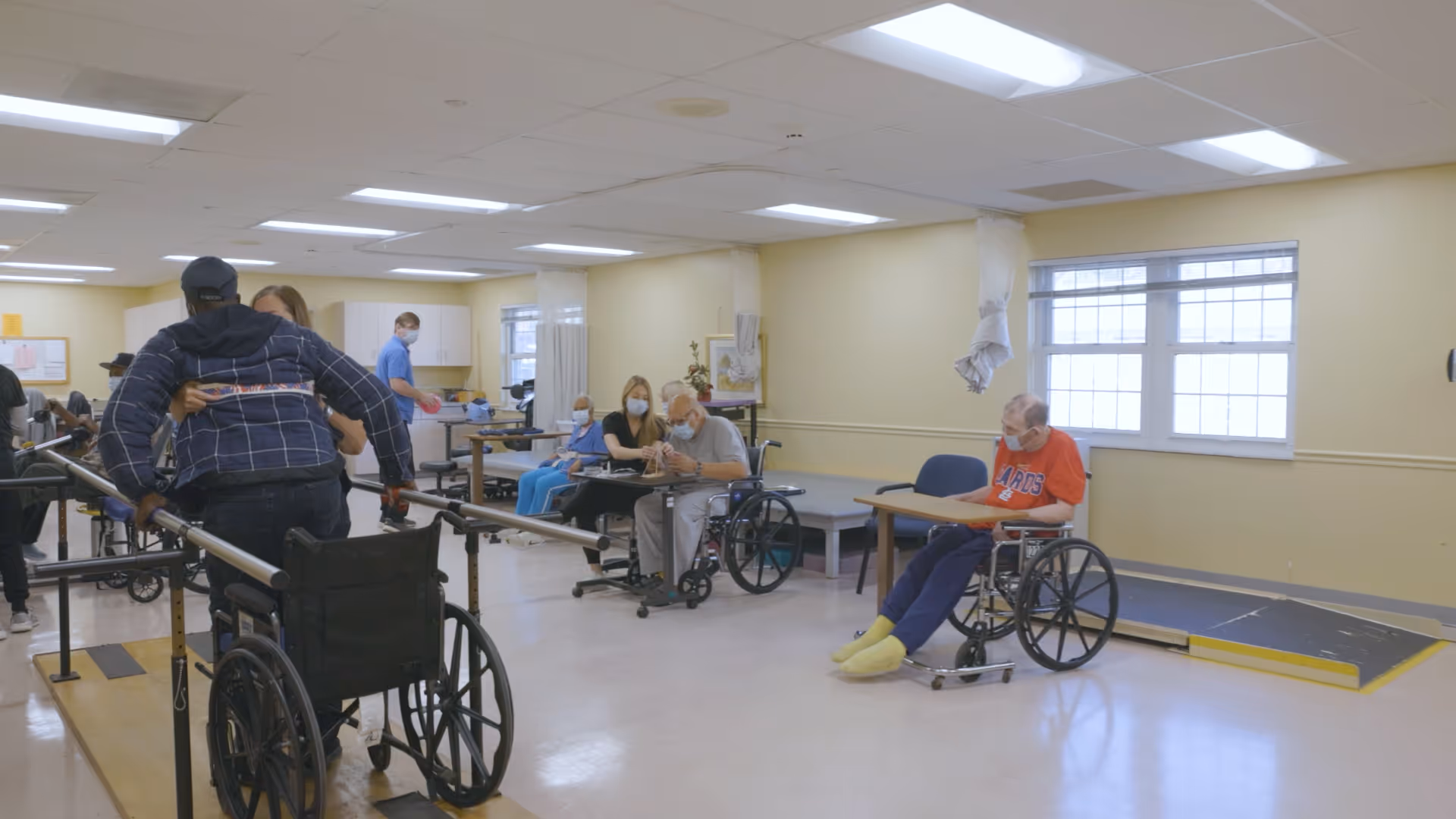 A rehabilitation room in a senior living facility where elderly individuals in wheelchairs are engaged in physical therapy activities. A staff member assists a person practicing walking with parallel bars. Other residents and staff are seated or standing in the background, some wearing masks. The room has large windows, light yellow walls, and a ramp along one side.