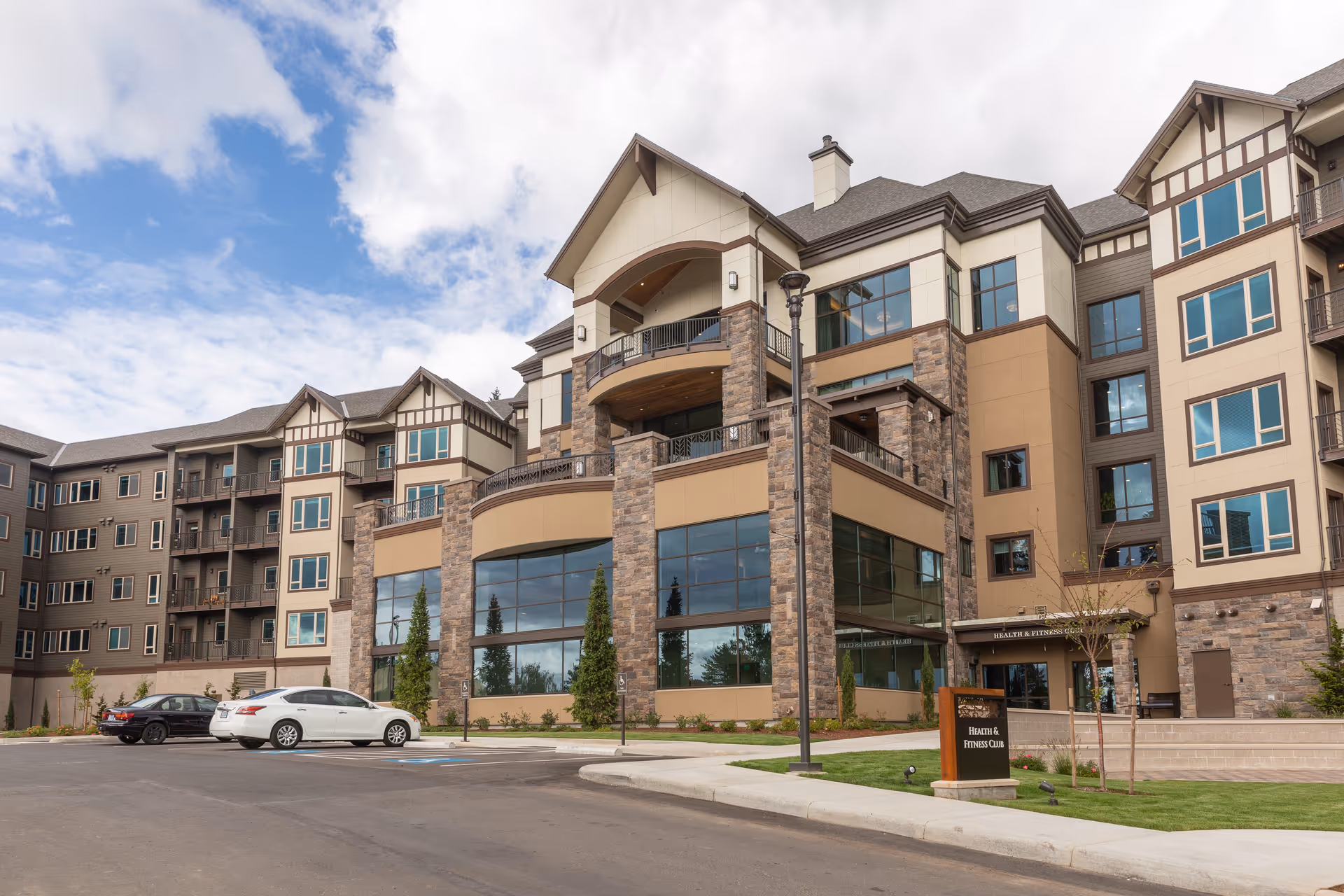Large multi-story senior living building with stone and stucco facade, balconies and parked cars in front under a partly cloudy sky.