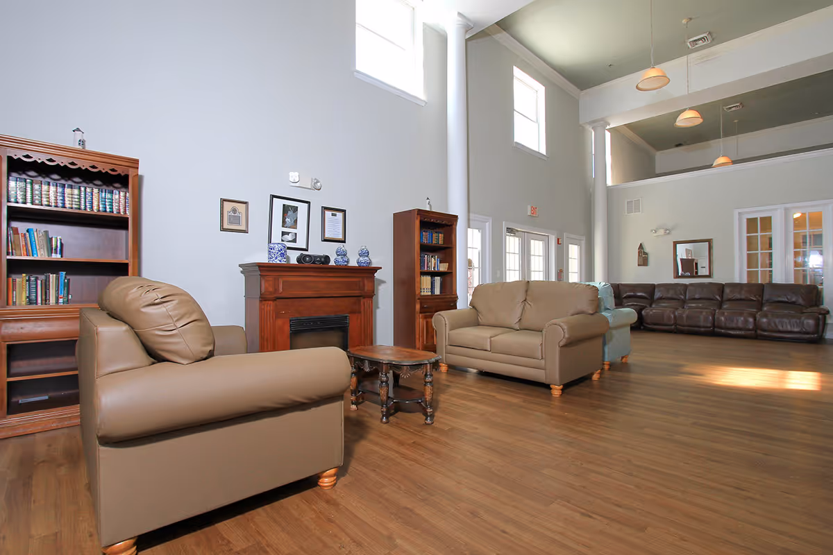 A spacious assisted living facility common area with high ceilings and large windows. The room features multiple seating options including a tan armchair, a tan loveseat, and a dark brown leather sofa. There are two wooden bookshelves filled with books and decorative items, and a wooden fireplace mantel with framed pictures and blue and white ceramic vases. The floor is wood, and pendant lights hang from the ceiling.
