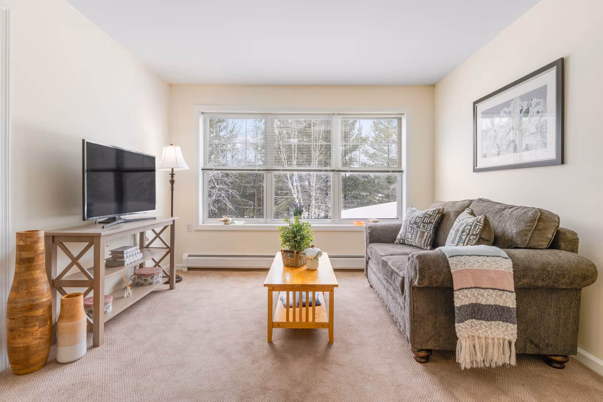 A cozy living room with a large window showing trees outside. The room features a gray sofa with patterned pillows and a striped throw blanket, a wooden coffee table with plants on it, a TV on a wooden stand, a floor lamp, and decorative vases on the floor.