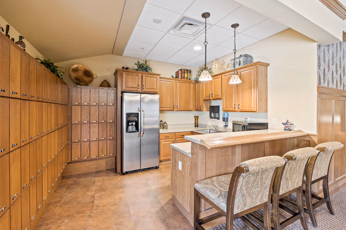 A kitchen area with wooden cabinets, a stainless steel refrigerator, a countertop with a sink, and three upholstered bar stools. The kitchen has pendant lights hanging from the ceiling and decorative items on top of the cabinets. There are also multiple wooden lockers or storage compartments along one wall.