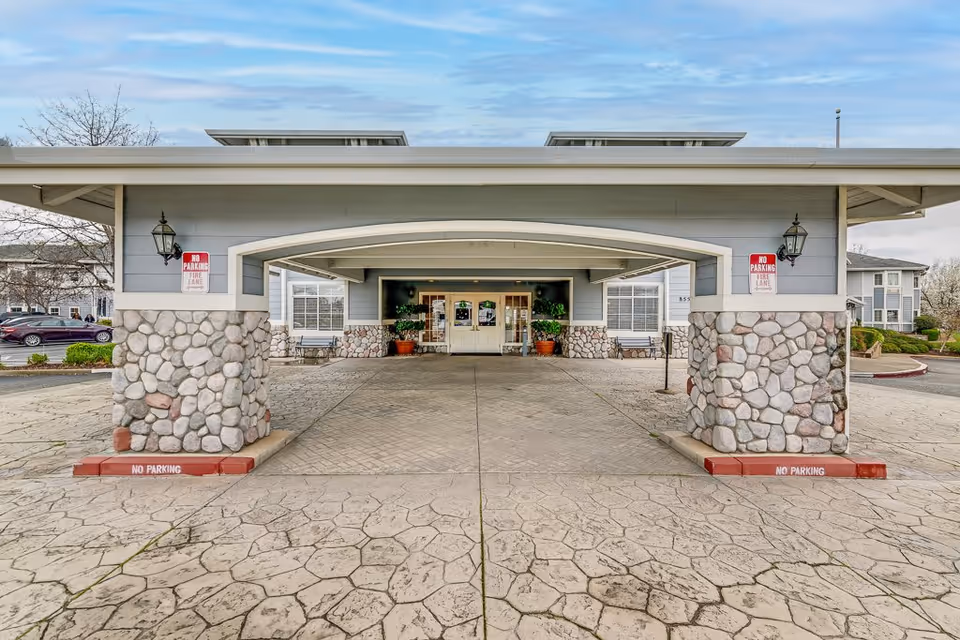 Entrance area of a senior living facility with a covered drop-off zone supported by stone pillars. The building has light gray siding with white trim and two lantern-style lights mounted on the pillars. There are 'No Parking Fire Lane' signs on the pillars and red curbs marked 'No Parking'. Two benches and potted plants are visible near the entrance doors.