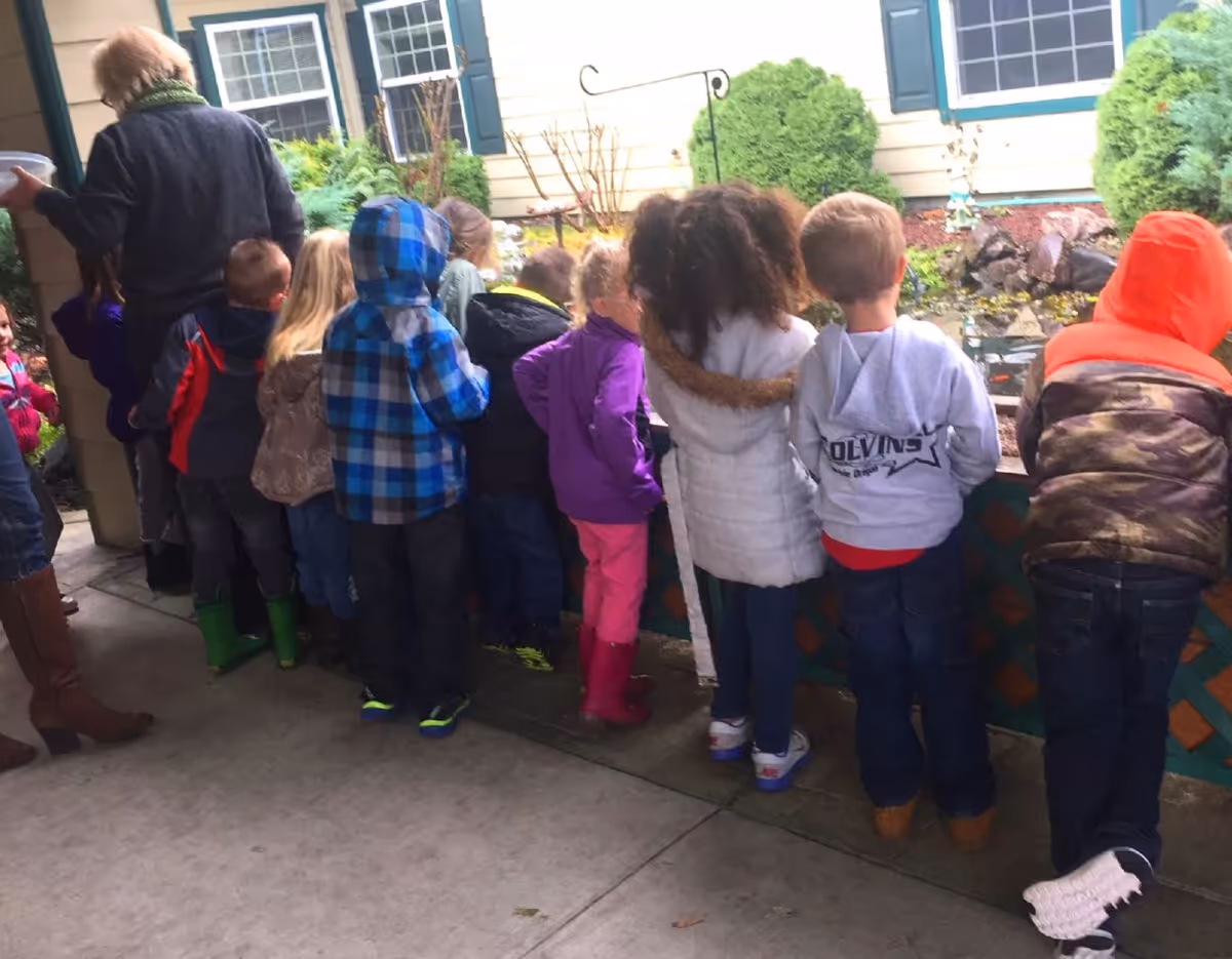 A group of children dressed in colorful jackets and boots stand in a row facing a small garden pond outside a building with windows and green shutters. An adult stands nearby, partially visible, wearing brown boots and a dark sweater.