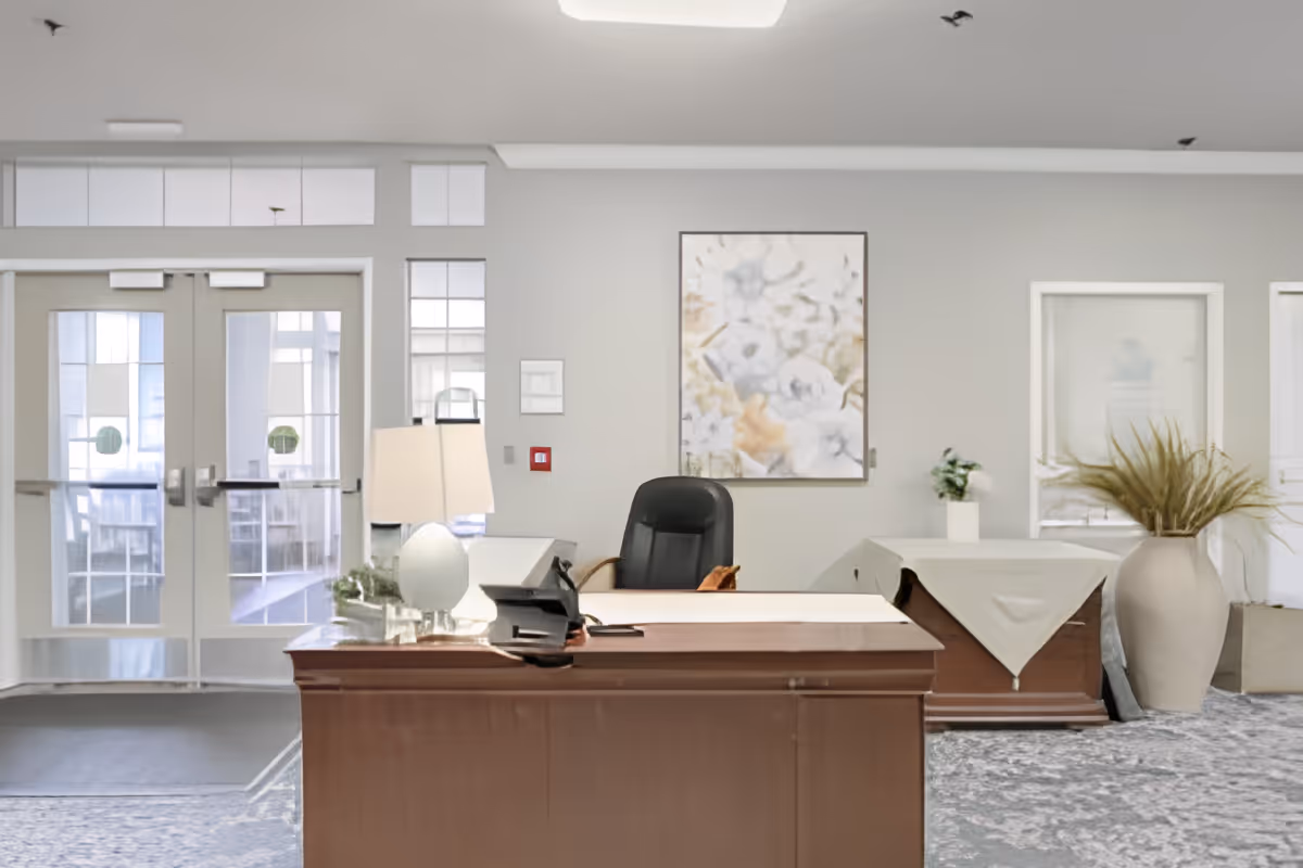 Reception area with a wooden desk, a black office chair, a table lamp, and a phone. Behind the desk is a wall with a floral painting, two windows, and a large vase with dried plants. Double glass doors lead to another room with tables and chairs.