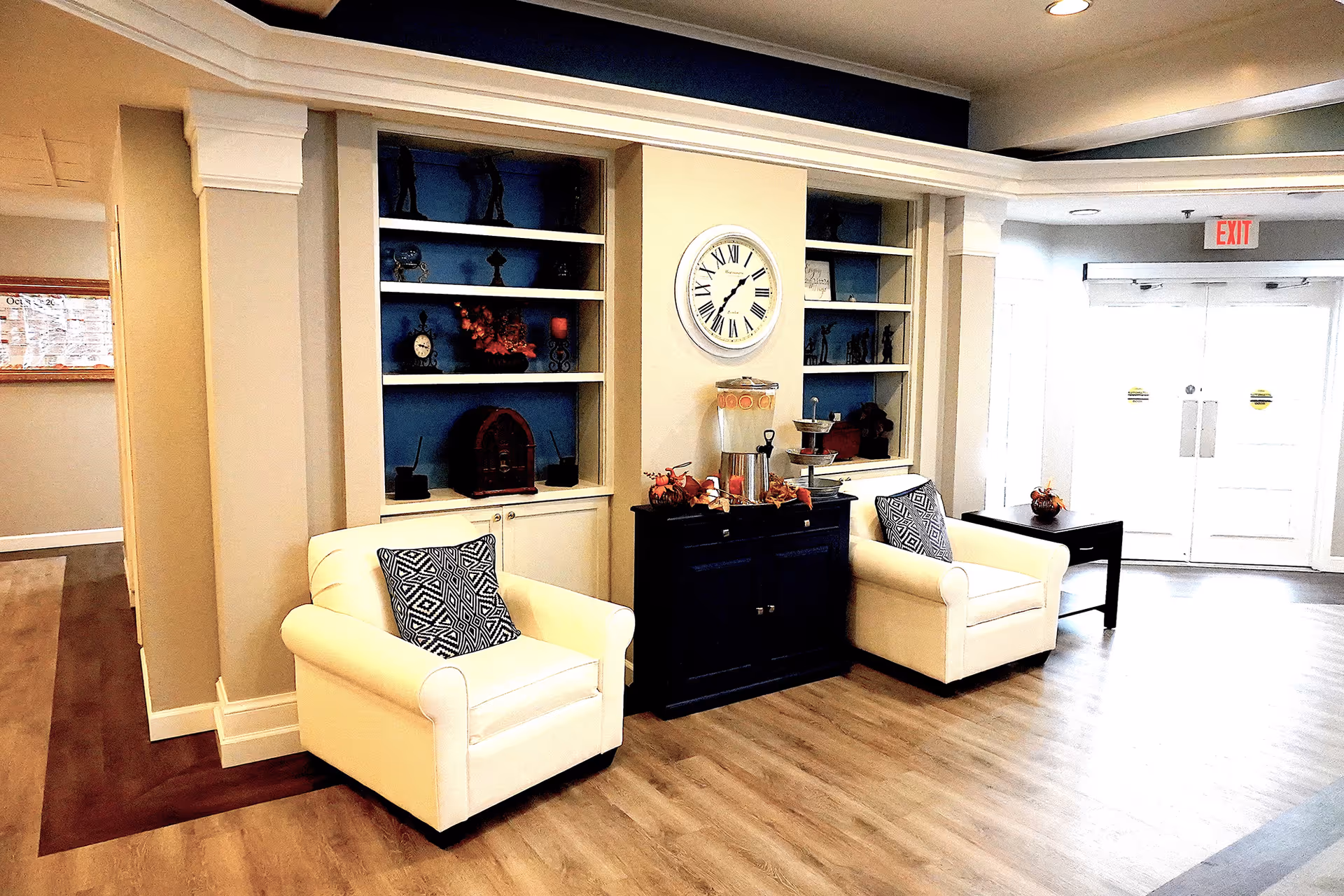 A cozy seating area in a senior living facility with two white armchairs, each with a patterned cushion. Between the chairs is a dark wooden cabinet with a beverage dispenser and decorative items on top. Behind the cabinet is a built-in shelving unit with various decorative objects and a large wall clock. The area has wood flooring and is well-lit with natural light coming from double glass doors in the background.