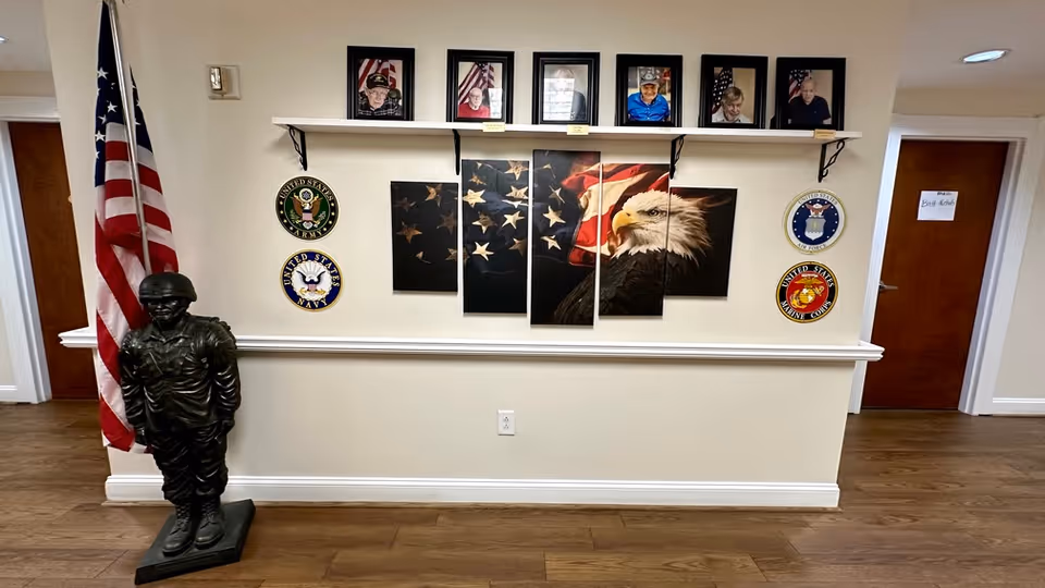 Interior wall display in a senior living facility featuring a large American flag and bald eagle artwork divided into five panels. Above the artwork are six framed photos of veterans. On either side of the artwork are four circular emblems representing branches of the United States military: Army, Navy, Air Force, and Marine Corps. To the left of the display is a statue of a soldier standing next to an American flag. Two doors are visible on either side of the wall, one with a sign reading 'Bath Robe'.