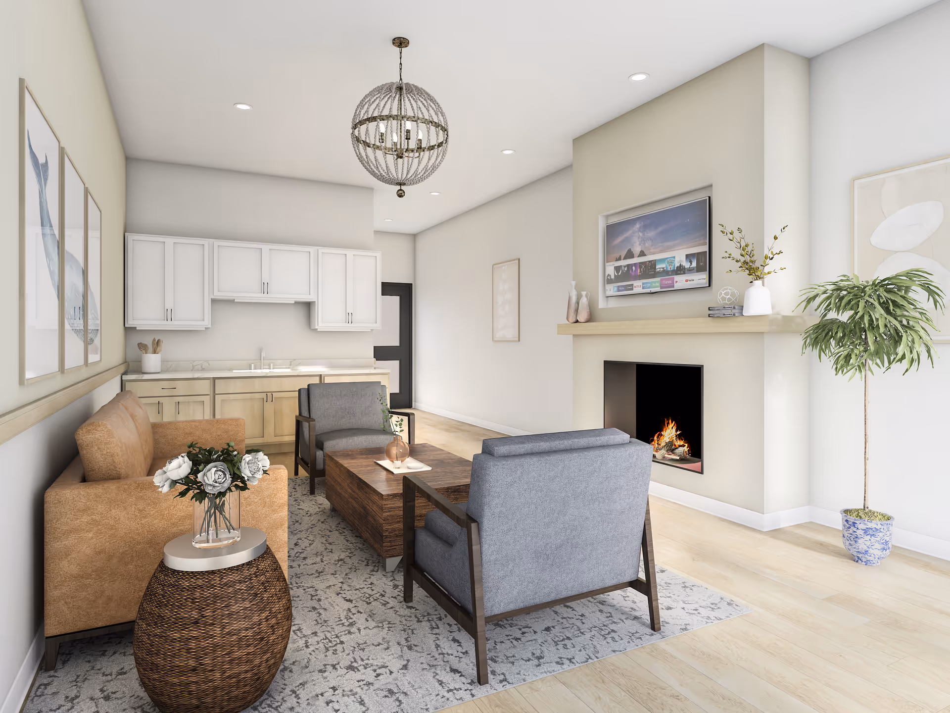 A bright and modern living room area featuring a brown sofa, two gray armchairs, a wooden coffee table, and a round wicker side table with a vase of flowers. The room has light wood flooring, a patterned area rug, a fireplace with a flat-screen TV mounted above it, and a tall potted plant in a blue and white ceramic pot. The back wall includes a kitchenette with white upper cabinets and light wood lower cabinets. The walls are decorated with framed artwork.