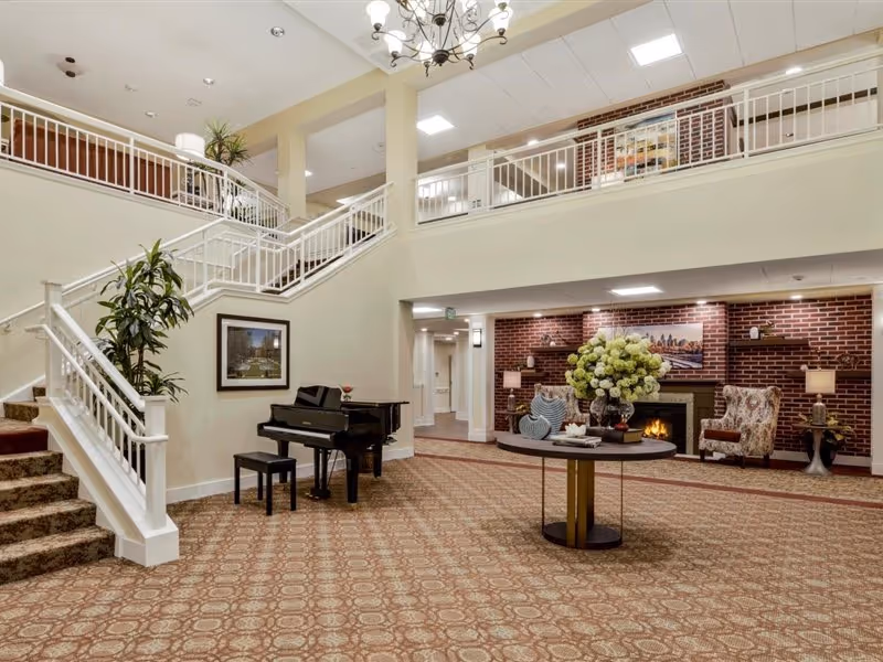 Spacious two-story lobby with a grand staircase, piano, central table with floral arrangement, and a seating area by a fireplace.