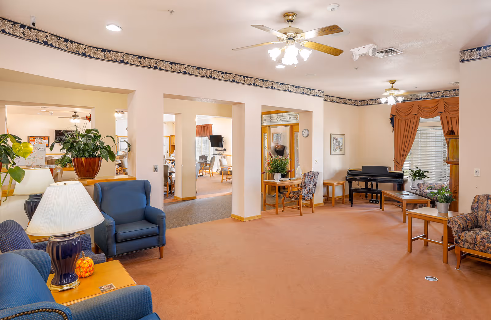 A spacious living room area in an assisted living facility with blue upholstered chairs, a table lamp, potted plants, a piano, floral curtains, and ceiling fans with lights. The room has beige walls with a floral border near the ceiling and carpeted floors. There are open doorways leading to other rooms with tables and chairs visible.