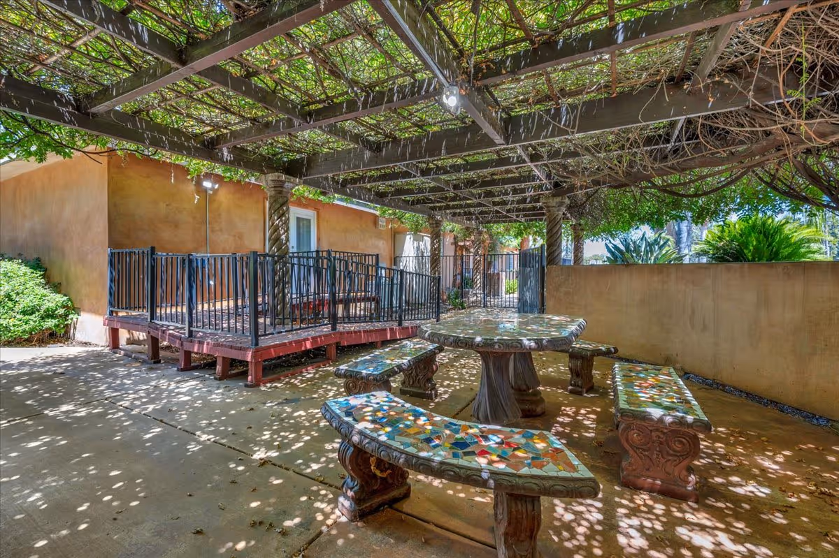 Outdoor patio area with a pergola covered in vines providing shade. Underneath, there is a mosaic-tiled stone table with matching curved benches. The patio is adjacent to a building with a beige stucco exterior and a small raised deck with black metal railings.
