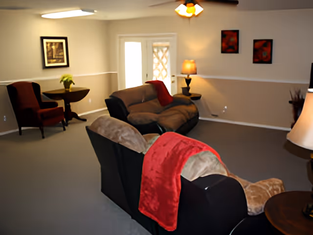 A cozy assisted living common room with sofas, armchairs, side tables, lamps, and wall art.