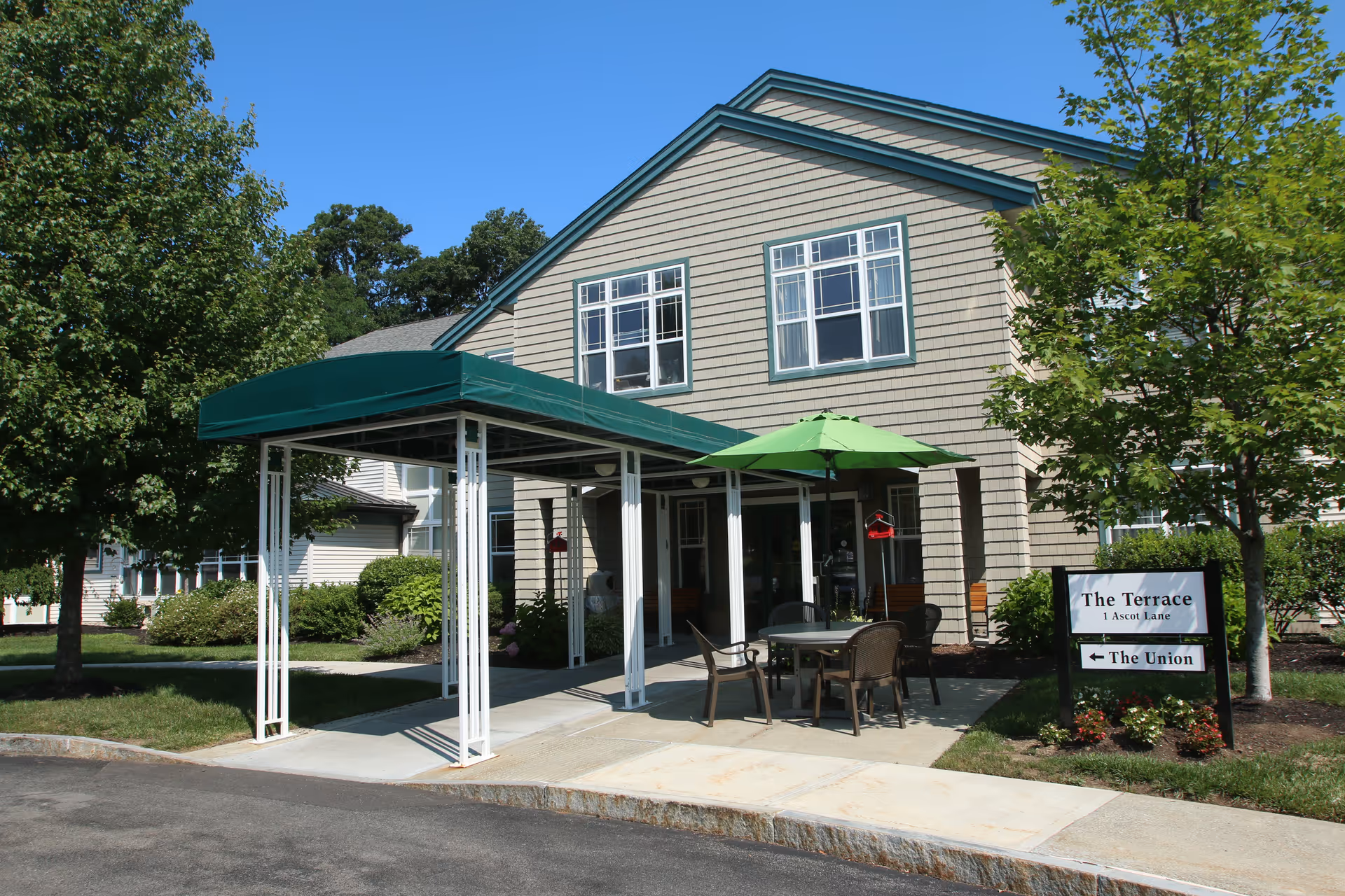 Exterior view of a senior living facility building with beige siding and green trim. There is a covered entrance with a green canopy and an outdoor seating area with a table, chairs, and a green umbrella. Trees and shrubs surround the building, and a sign near the entrance reads 'The Terrace 1 Ascot Lane' with an arrow pointing to 'The Union'.