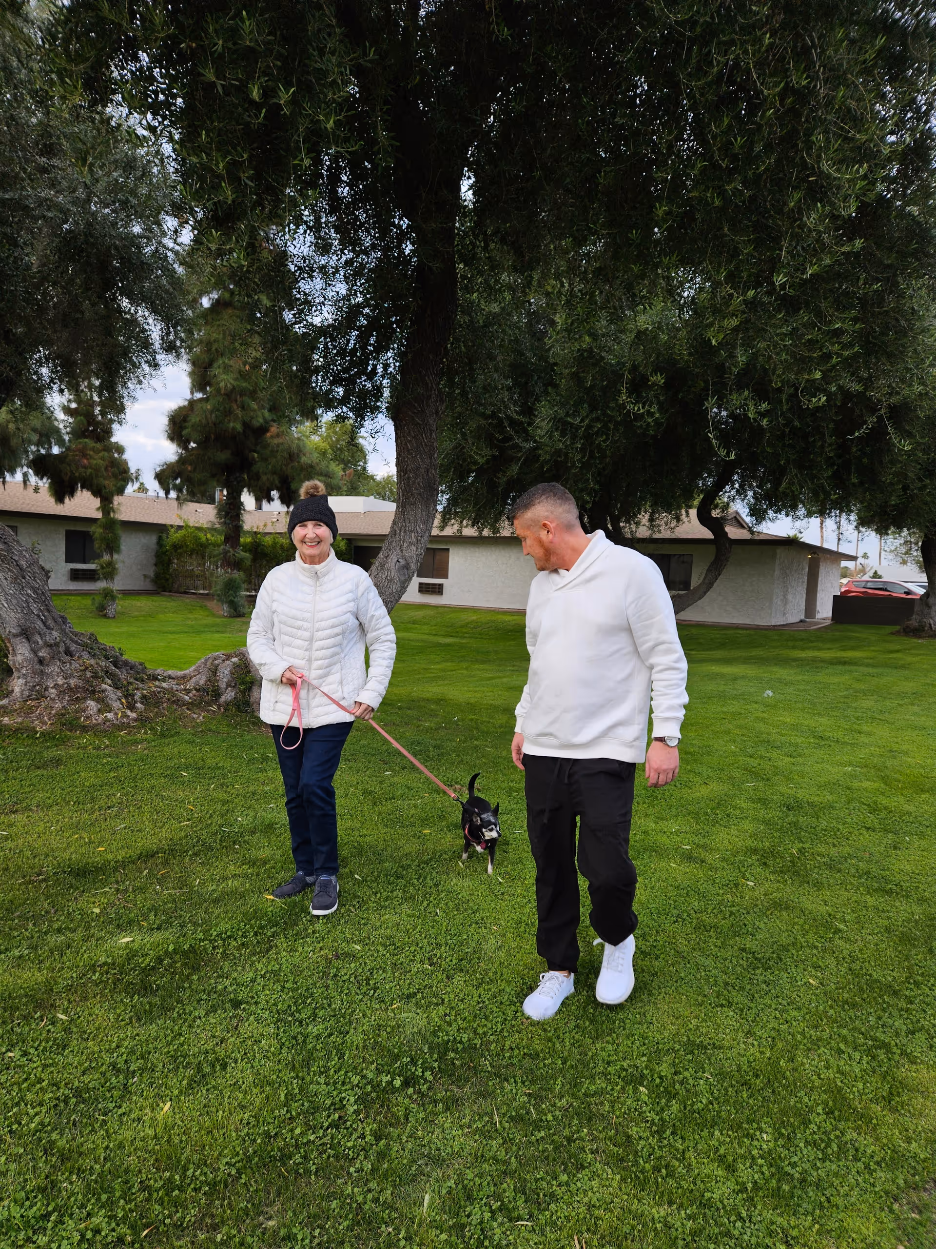 An elderly woman and a man walking a small black and white dog on a leash in a grassy outdoor area with large trees and a single-story building in the background. The woman is wearing a white jacket and black hat, and the man is dressed in a white sweater and black pants.