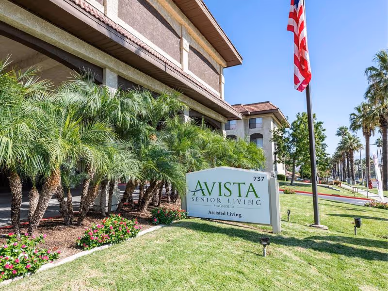 Exterior view of Vista Senior Living facility with a sign reading 'Vista Senior Living Magnolia Assisted Living' on a well-maintained lawn with palm trees and an American flag on a flagpole.