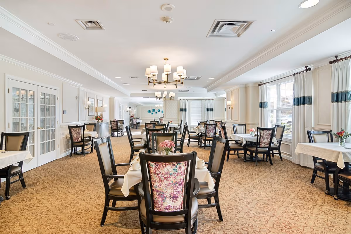A bright and spacious dining room with multiple round tables covered with white tablecloths. Each table is surrounded by dark wooden chairs with floral patterned backs. The room has beige walls, large windows with white and blue curtains, and elegant chandeliers hanging from the ceiling. There are small flower arrangements on each table.