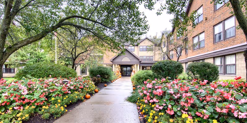 Pathway leading to the entrance of a brick senior living facility building, flanked by lush green bushes and colorful flower beds with pink, yellow, and red flowers. Trees with green leaves frame the scene on both sides.