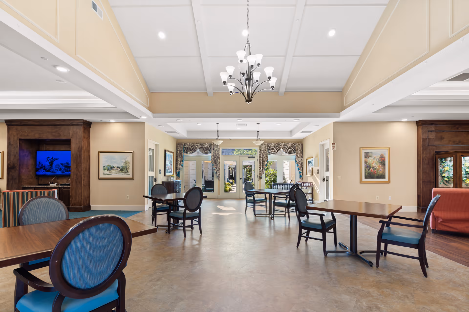 A spacious and well-lit common area in a senior living facility with several wooden tables and blue cushioned chairs arranged around the room. The room features a high ceiling with a chandelier, large windows with decorative curtains, framed artwork on the walls, and a television mounted in a wooden entertainment unit. There is a door leading to an outdoor area with greenery visible through the glass.