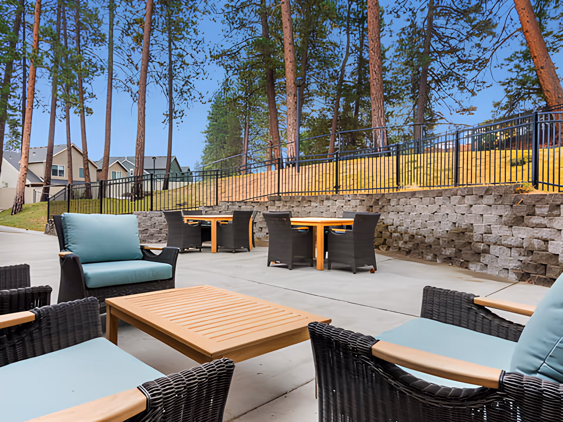 Outdoor patio area with several black wicker chairs featuring light blue cushions and wooden armrests, surrounding wooden tables. The patio is paved with concrete and bordered by a stone retaining wall and black metal fence. Tall pine trees and residential houses are visible in the background under a clear blue sky.