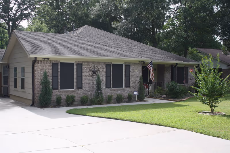 Single-story brick building with black shutters on the windows, a small porch with an American flag, and a neatly maintained lawn with shrubs and a small tree in front.