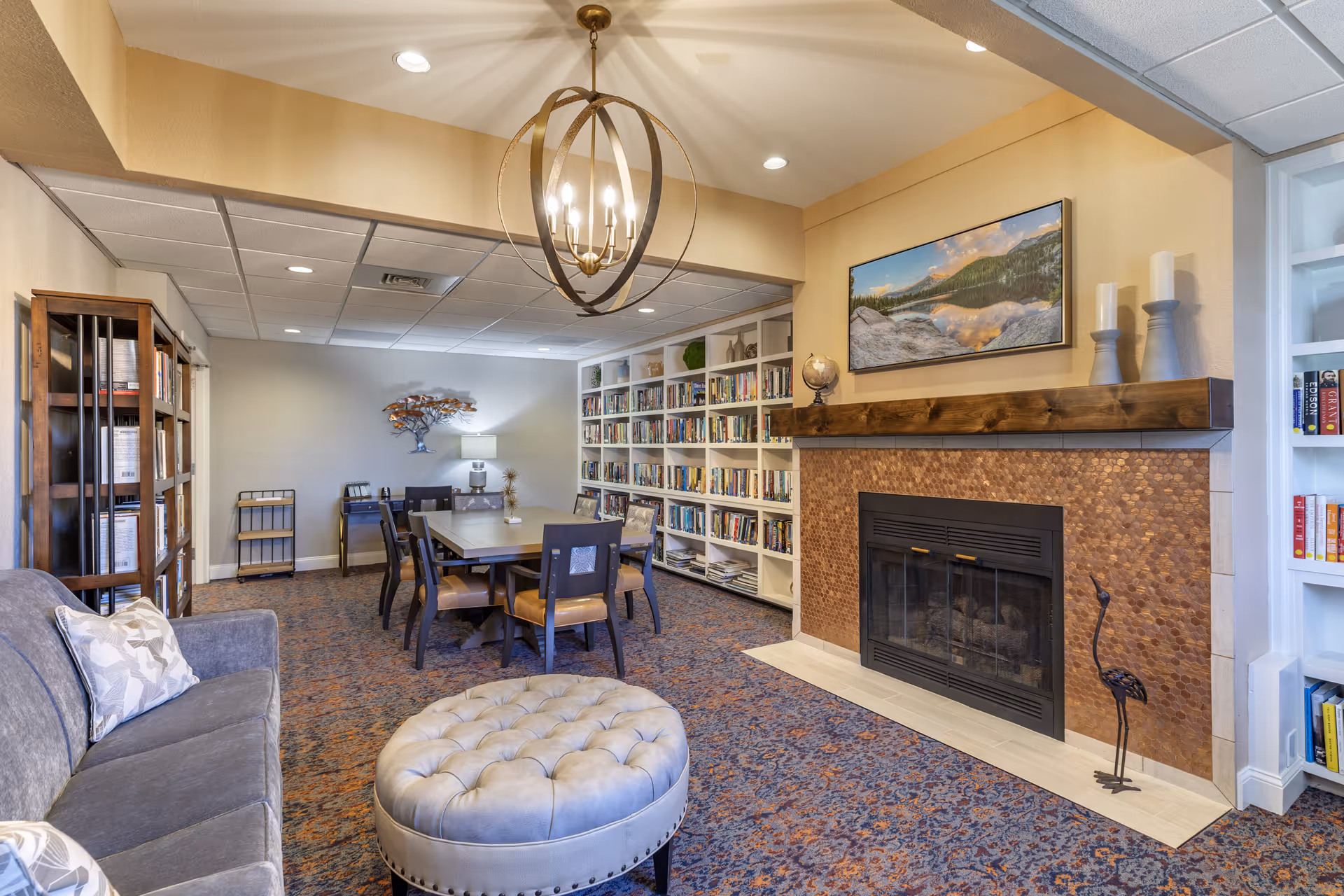 A cozy living room area in a senior living facility featuring a gray sofa with patterned pillows, a round tufted ottoman, a wooden dining table with chairs, built-in bookshelves filled with books, a fireplace with a wooden mantel decorated with candles and a globe, and a landscape painting above the fireplace. The room has a patterned carpet and a modern chandelier hanging from the ceiling.