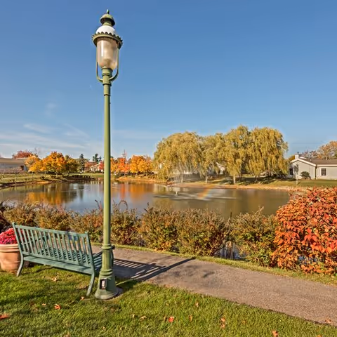 A peaceful outdoor scene at Elison Independent Living of Orchard Glen featuring a green bench and a vintage-style street lamp beside a paved walkway. In the background, there is a pond with a fountain, surrounded by autumn-colored trees and shrubs under a clear blue sky.