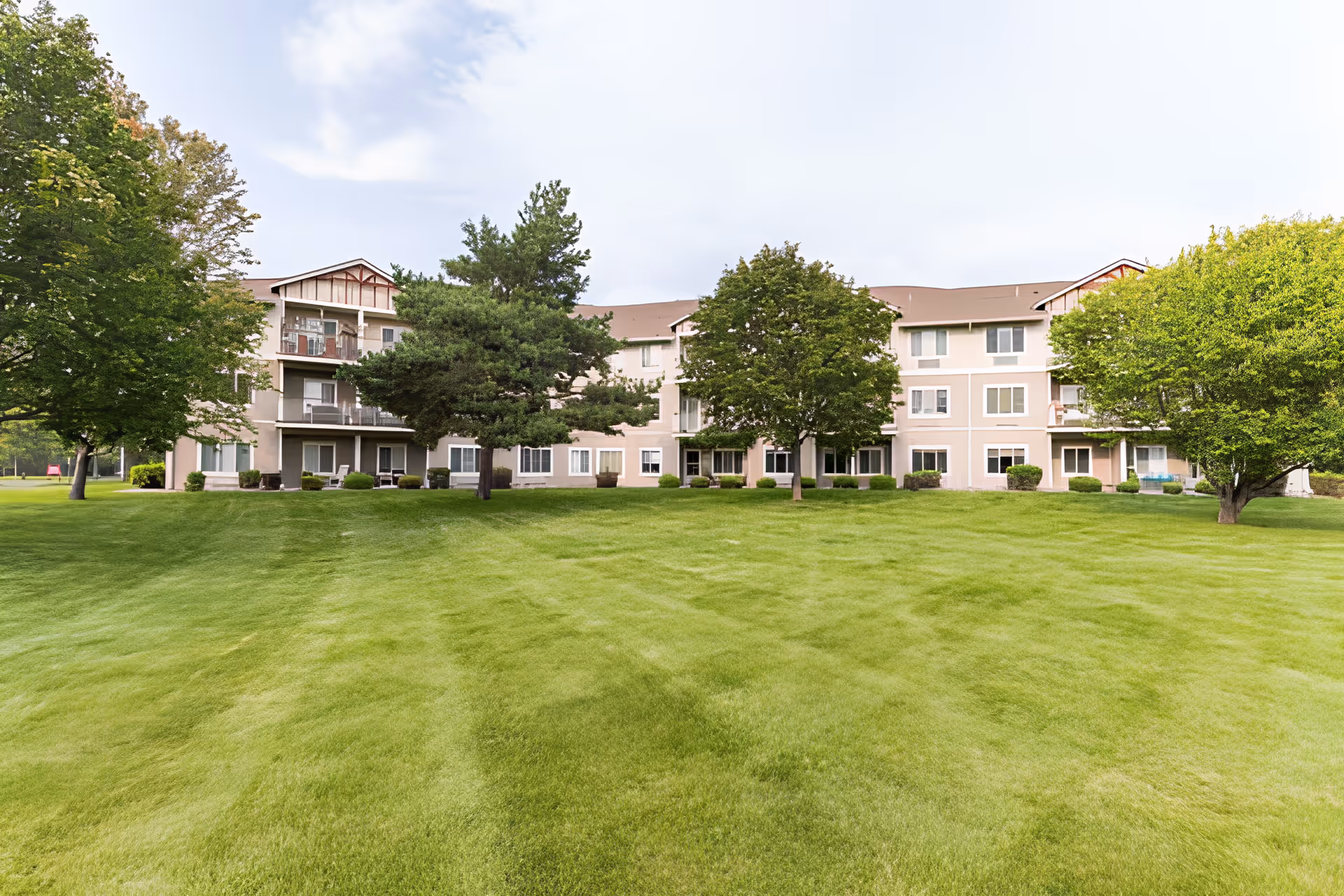 Three-story residential building with balconies and windows set behind a wide mowed lawn and scattered trees.