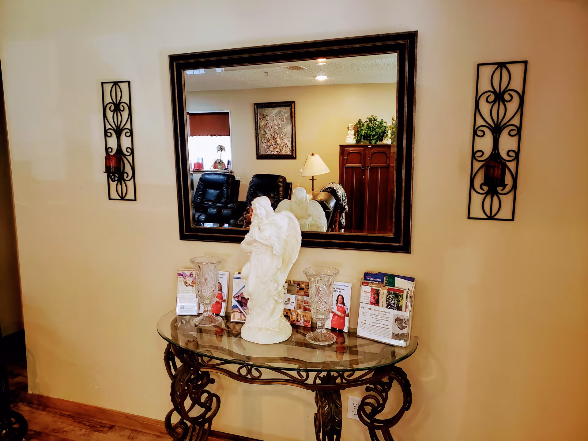 A decorative glass table with ornate black metal legs against a beige wall. On the table are two crystal vases, a white angel statue, and several brochures. Above the table is a large rectangular mirror reflecting a room with black leather chairs, a lamp, a wooden cabinet, and a framed picture. On either side of the mirror are black wrought iron wall sconces with red candles.