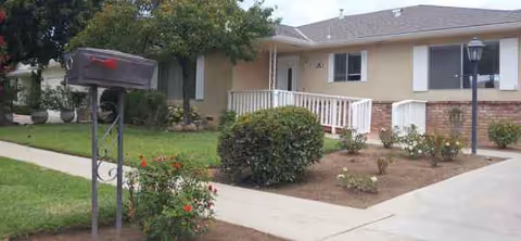 Front view of a senior living facility with walkway, shrubs, mailbox, and railing.