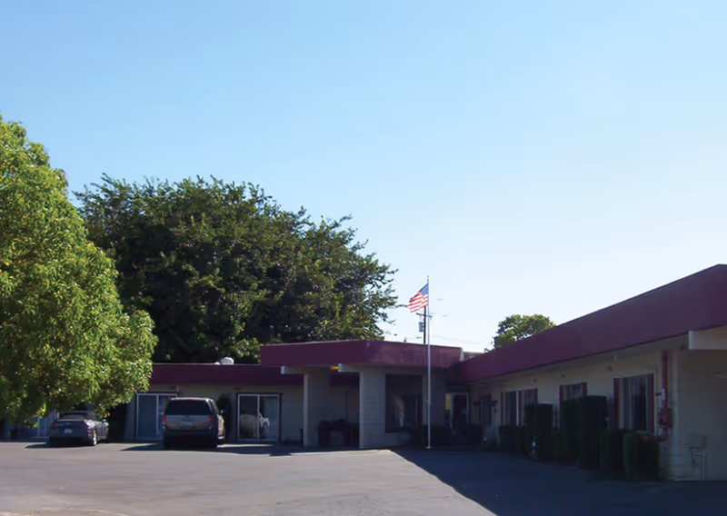 Exterior view of a single-story building with a maroon roof and beige walls, surrounded by trees and a paved parking area with two parked cars. An American flag is flying on a flagpole near the entrance.