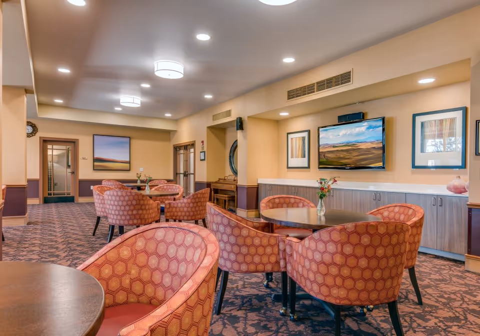 Comfortable communal lounge/dining area with round tables, patterned armchairs, a wall-mounted TV, and framed artwork.