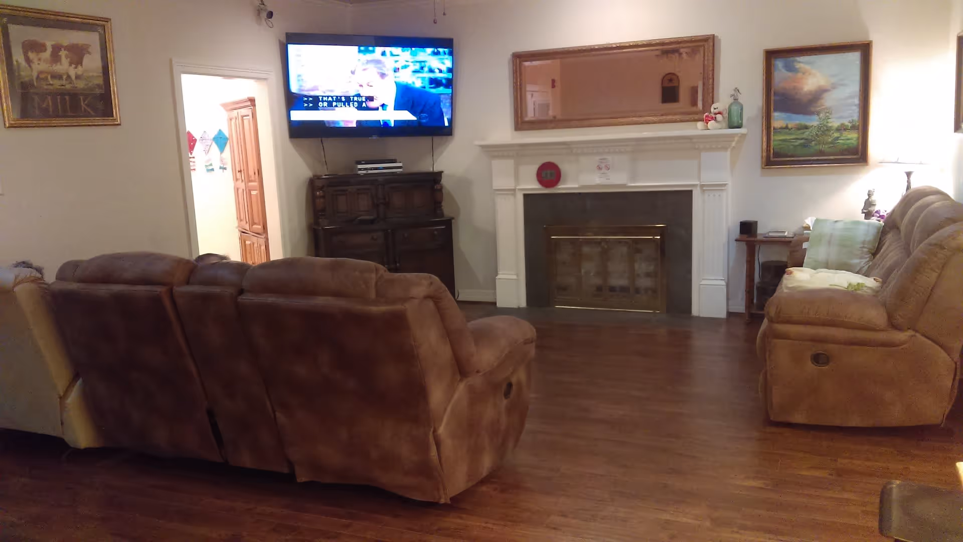 A cozy living room with brown reclining sofas facing a wall-mounted flat-screen TV above a wooden cabinet. There is a white fireplace with a large mirror above it and framed paintings on the walls. The room has wooden flooring and soft lighting from a lamp on a side table.