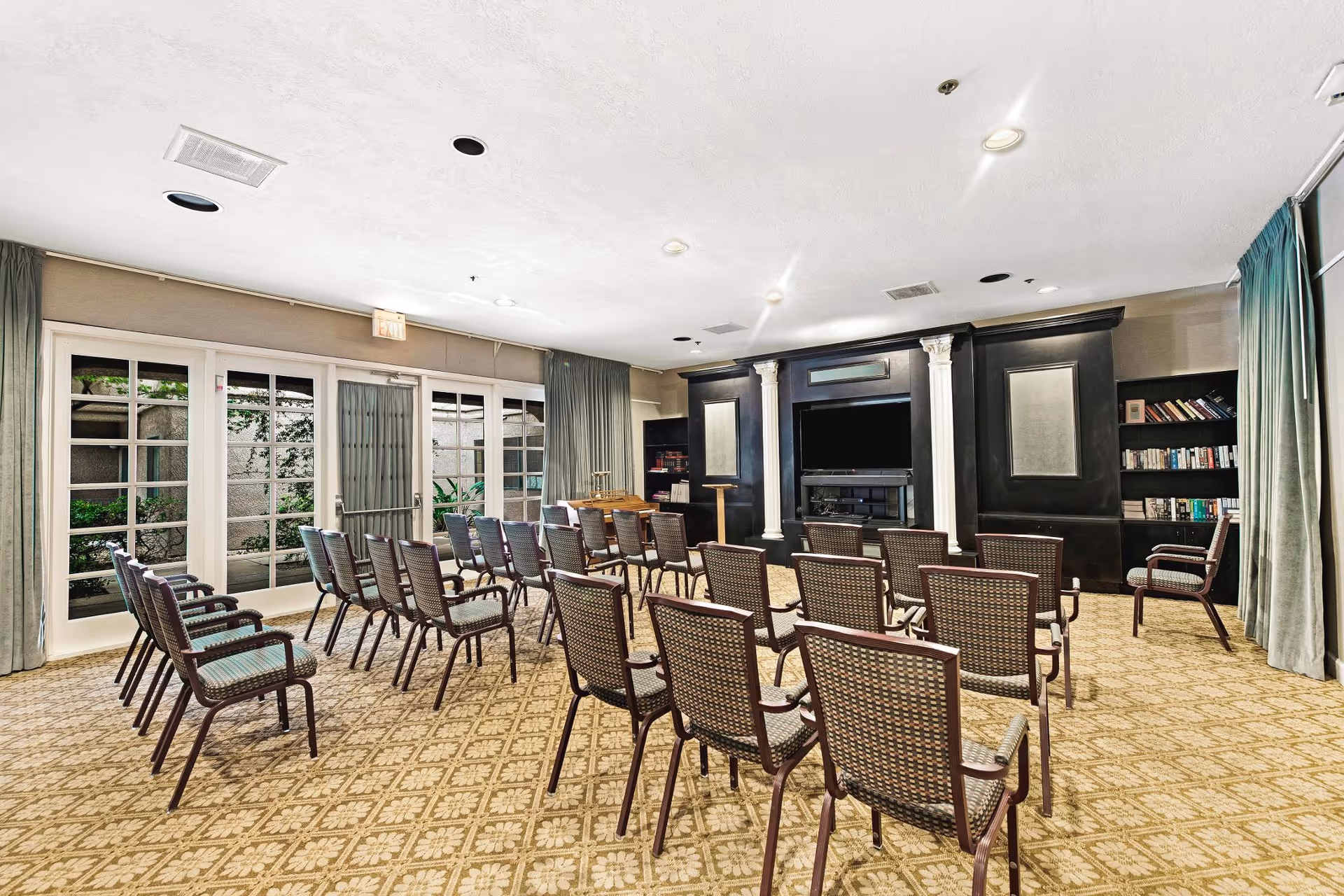 A meeting or presentation room with rows of brown chairs facing a black entertainment center with a large flat-screen TV and built-in shelves filled with books. The room has beige patterned carpet, large windows with curtains on the left side, and a glass door leading outside.