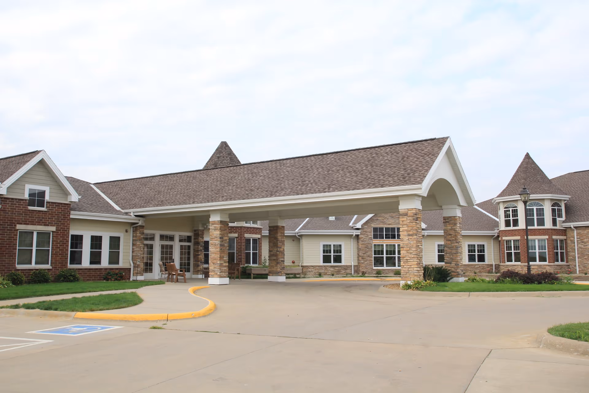 Front exterior view of a senior living facility named Highland Ridge with a covered entrance supported by stone pillars, brick and siding walls, multiple windows, and a driveway with a handicapped parking space.