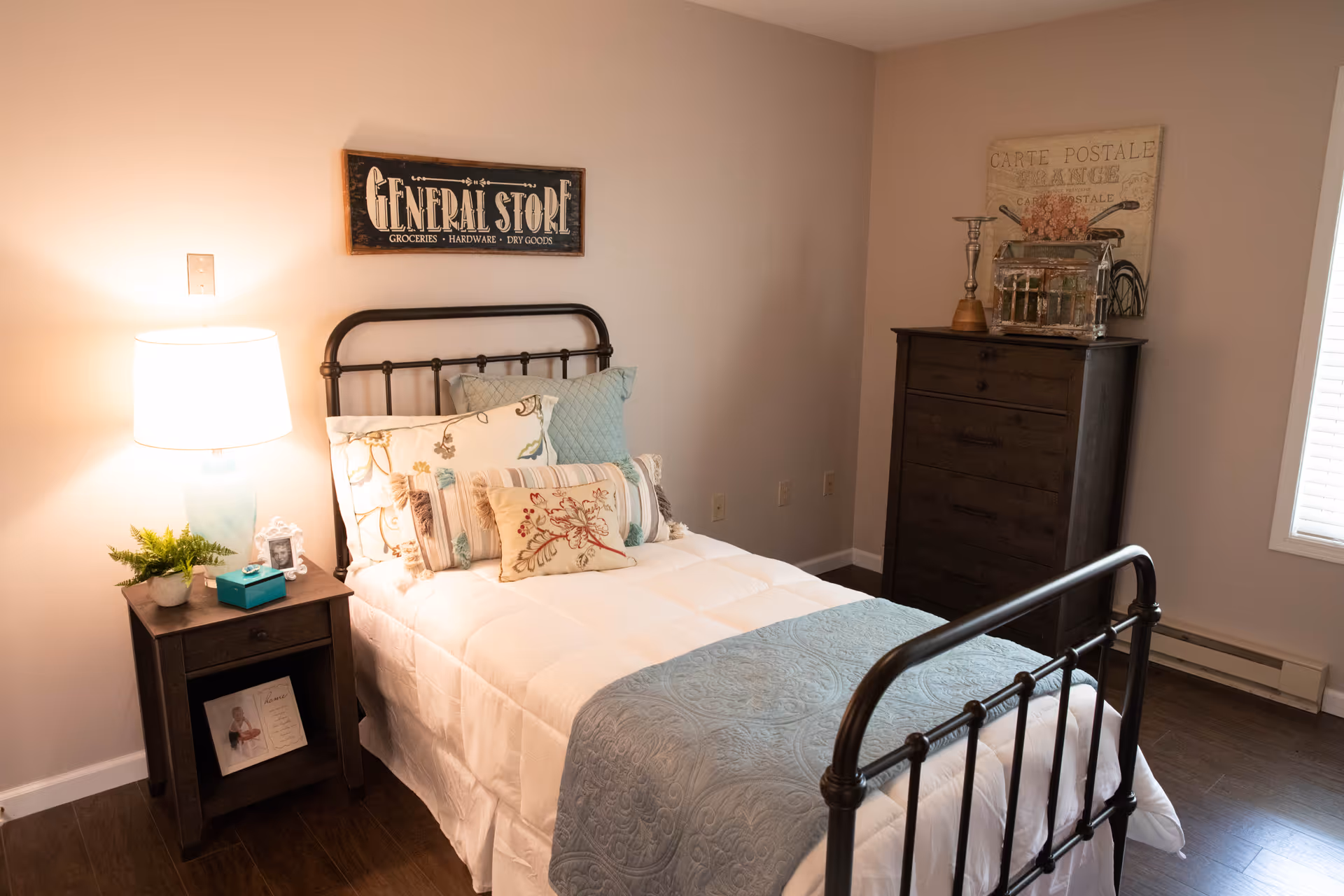 A cozy bedroom with a metal frame bed dressed in white bedding and multiple decorative pillows. A wooden nightstand with a lamp, small plant, and picture frame is beside the bed. On the wall above the bed is a sign that reads 'GENERAL STORE Groceries Hardware Dry Goods.' A wooden dresser with decorative items on top is against the far wall, next to a window with blinds.