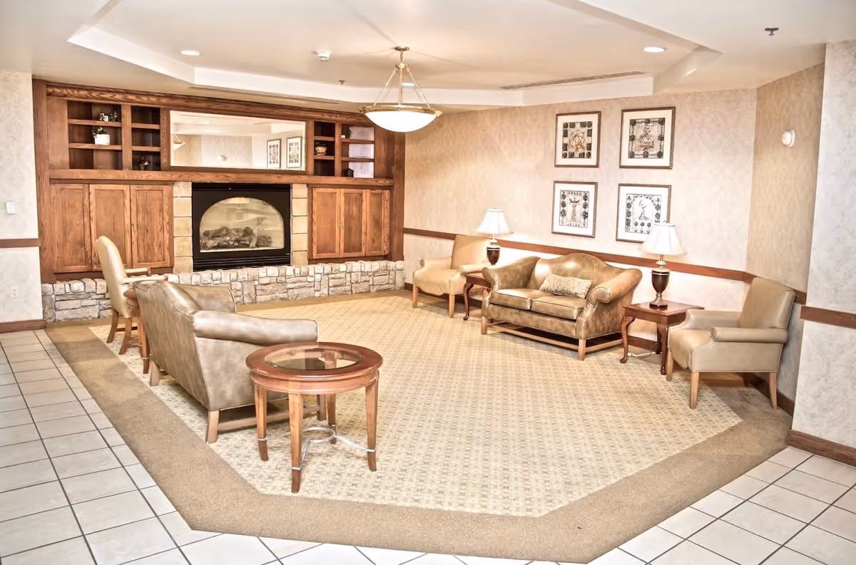 A cozy senior living room with beige walls and carpet, featuring a stone and wood fireplace with built-in shelves and cabinets. The room is furnished with a brown leather loveseat, two beige armchairs, a small wooden side table with a lamp, and a round glass-top coffee table. Four framed artworks hang on the wall above the loveseat, and a ceiling light fixture is centered above the seating area.