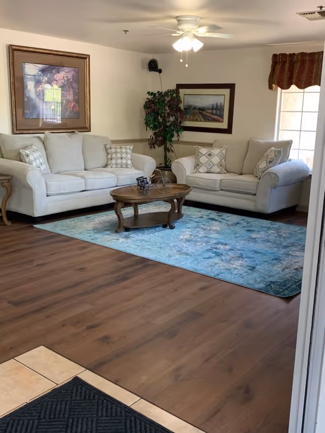 A cozy living room with two beige sofas adorned with patterned throw pillows, a wooden coffee table with a decorative bicycle sculpture, a blue area rug, a potted plant in the corner, framed artwork on the walls, and a ceiling fan with lights.