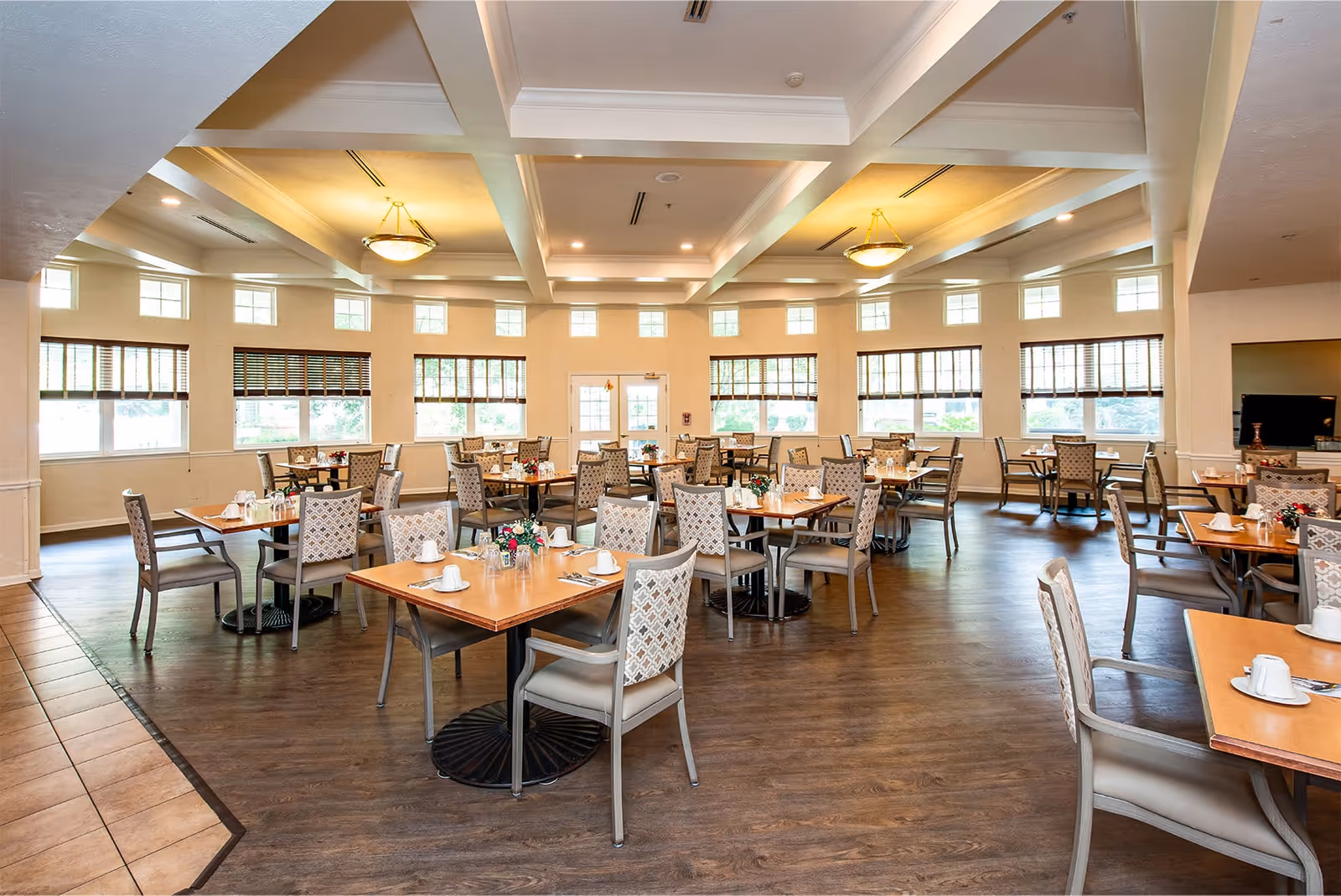 A spacious dining room with multiple wooden tables and cushioned chairs arranged neatly. Each table is set with white cups, saucers, and small flower arrangements. The room has large windows with blinds, allowing natural light to fill the space. The ceiling features recessed lighting and decorative beams.