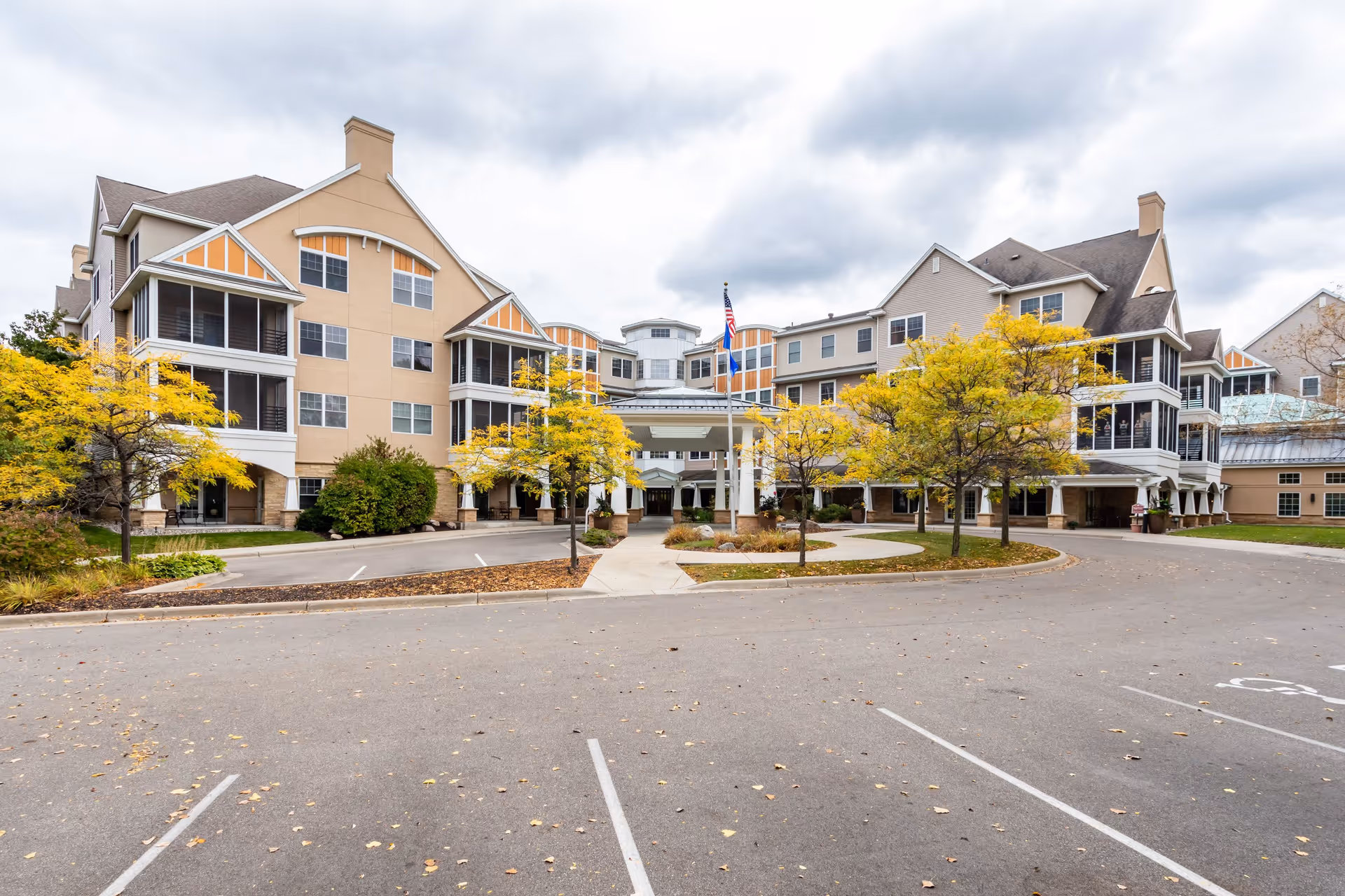 Exterior view of a large senior living facility building with beige and light brown walls, multiple windows, and screened balconies. The foreground shows a circular driveway with a flagpole displaying the American flag, surrounded by small trees with yellow autumn leaves. The sky is overcast.