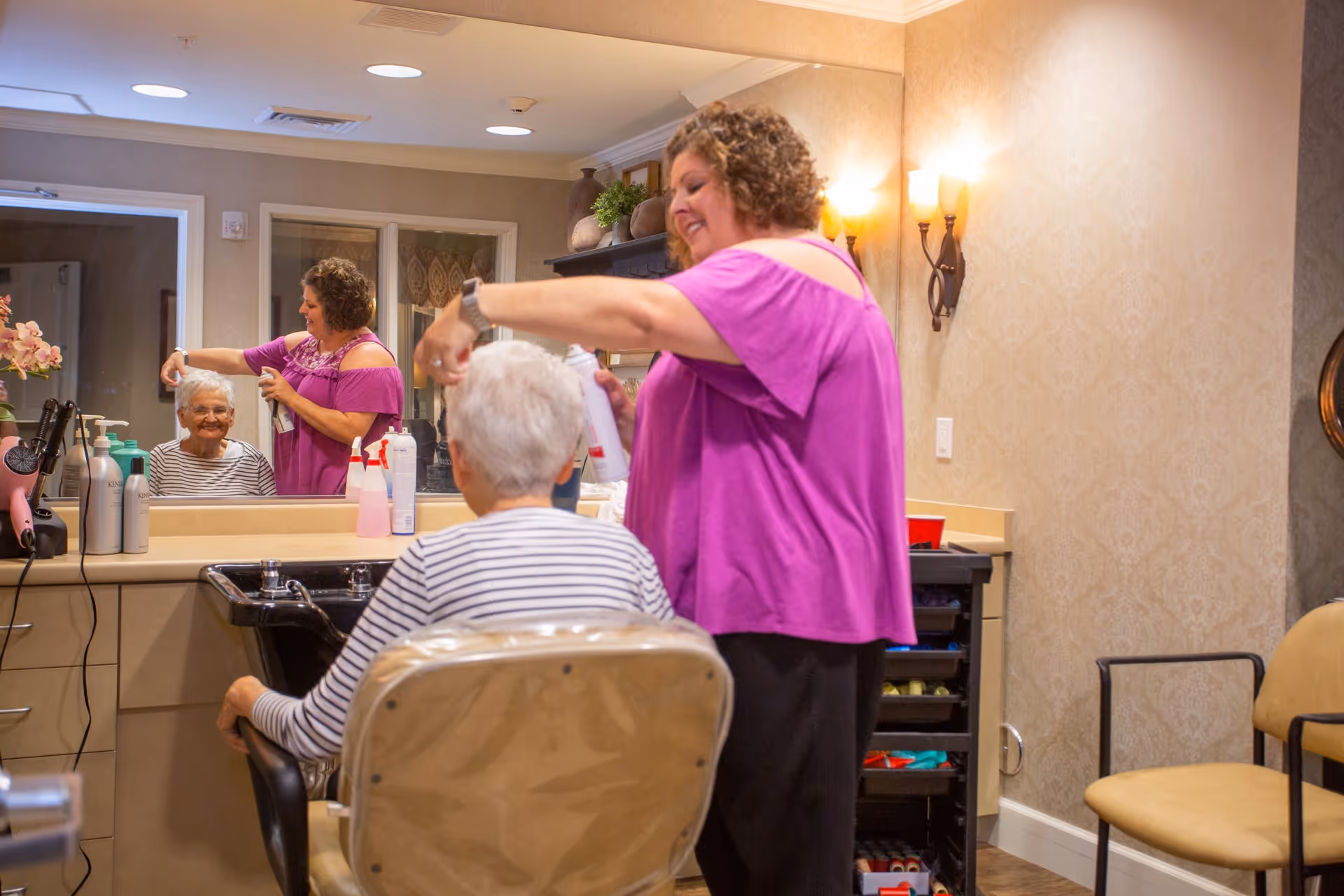A woman in a purple top is styling the hair of an elderly woman seated in a salon chair in front of a large mirror. The elderly woman is smiling and wearing a striped shirt. The room has beige walls with decorative sconces and various hair care products on the counter.