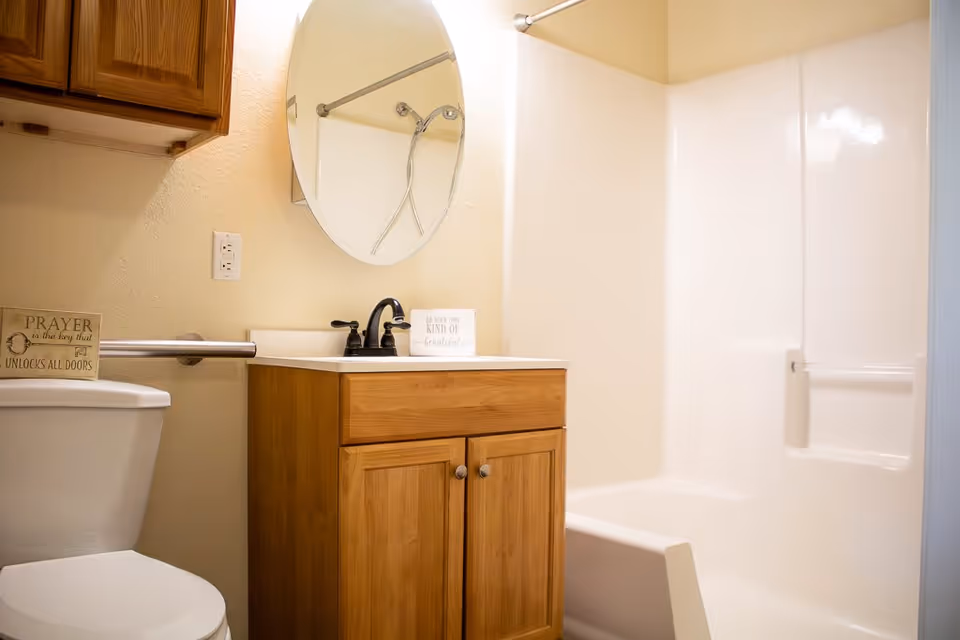 A bathroom with a white toilet, wooden vanity with a black faucet, an oval mirror above the sink, a shower with a white surround and a shower curtain rod, and a wooden cabinet mounted on the wall. There is a small sign on the toilet tank that reads 'PRAYER is the key that UNLOCKS ALL DOORS.'
