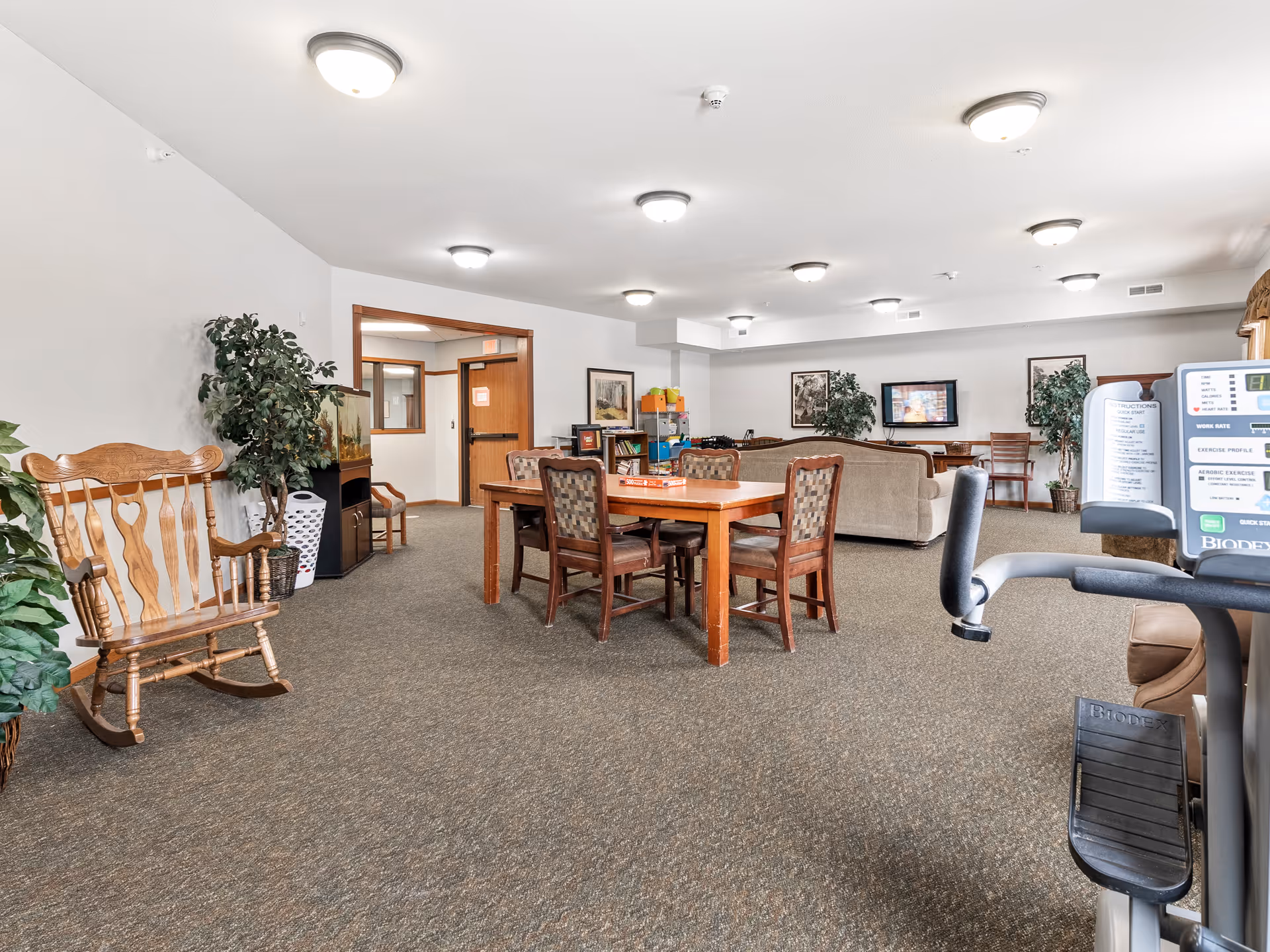 A spacious senior living common area with a wooden rocking chair, a large wooden table with four chairs, a beige couch, a flat-screen TV mounted on the wall, potted plants, and exercise equipment in the foreground. The room has carpeted floors, white walls, and multiple ceiling lights.