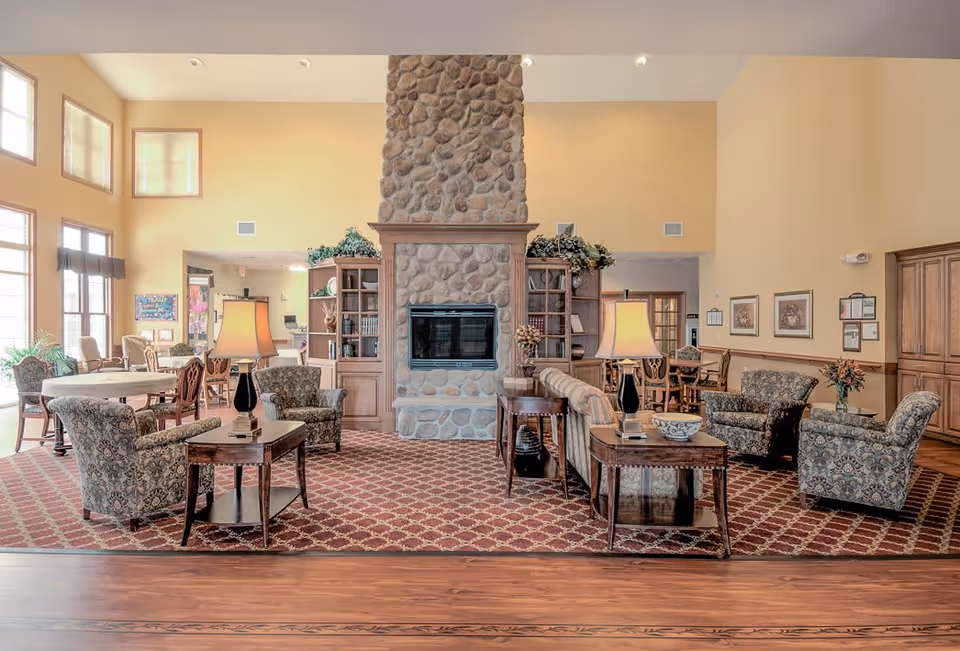 A spacious and well-lit living room area with a large stone fireplace in the center. The room features patterned armchairs, sofas, wooden side tables with lamps, and built-in wooden shelves flanking the fireplace. Large windows on the left allow natural light to fill the room, and there are additional tables and chairs in the background.