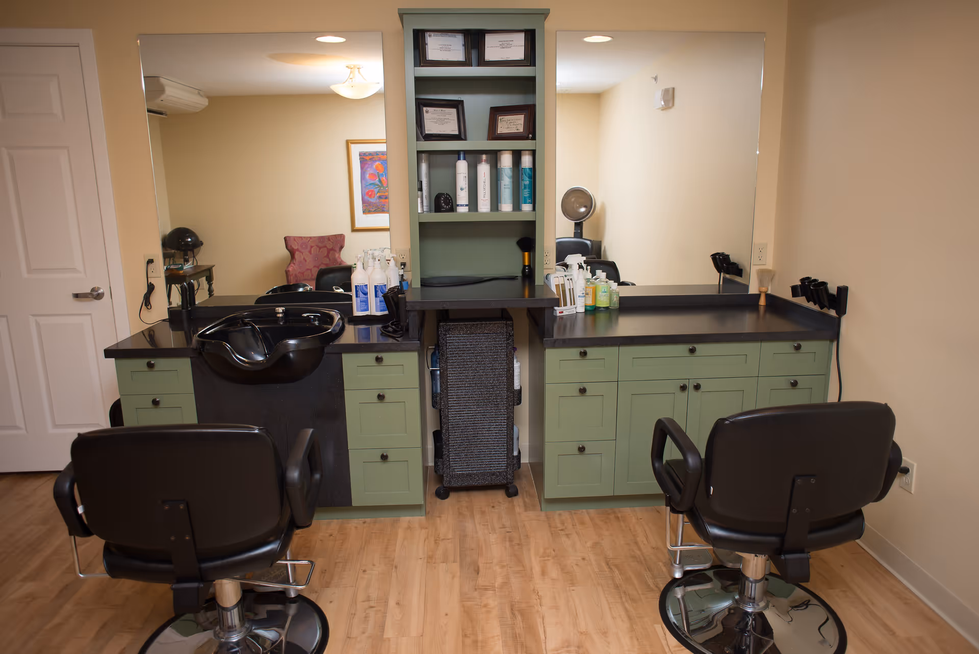 Interior view of a hair salon area with two black salon chairs facing large mirrors. There is a black hair washing sink on the left side, green cabinets with black countertops, and shelves with hair care products and framed certificates. The room has light-colored walls and wood flooring.